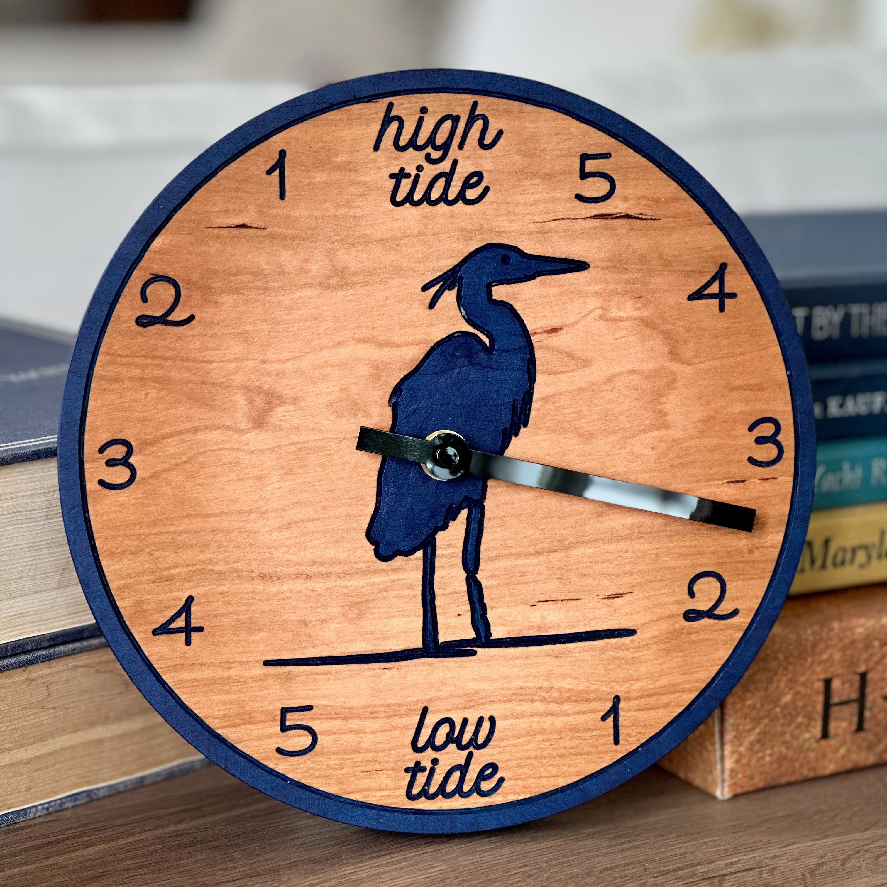 A round wooden wall clock with a navy blue border, a heron silhouette, and tide-themed text reading &