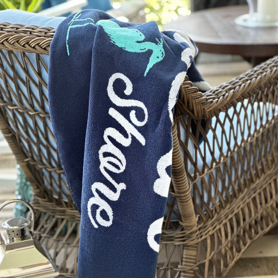 A navy blue towel draped over a wicker patio chair, featuring white cursive text reading &