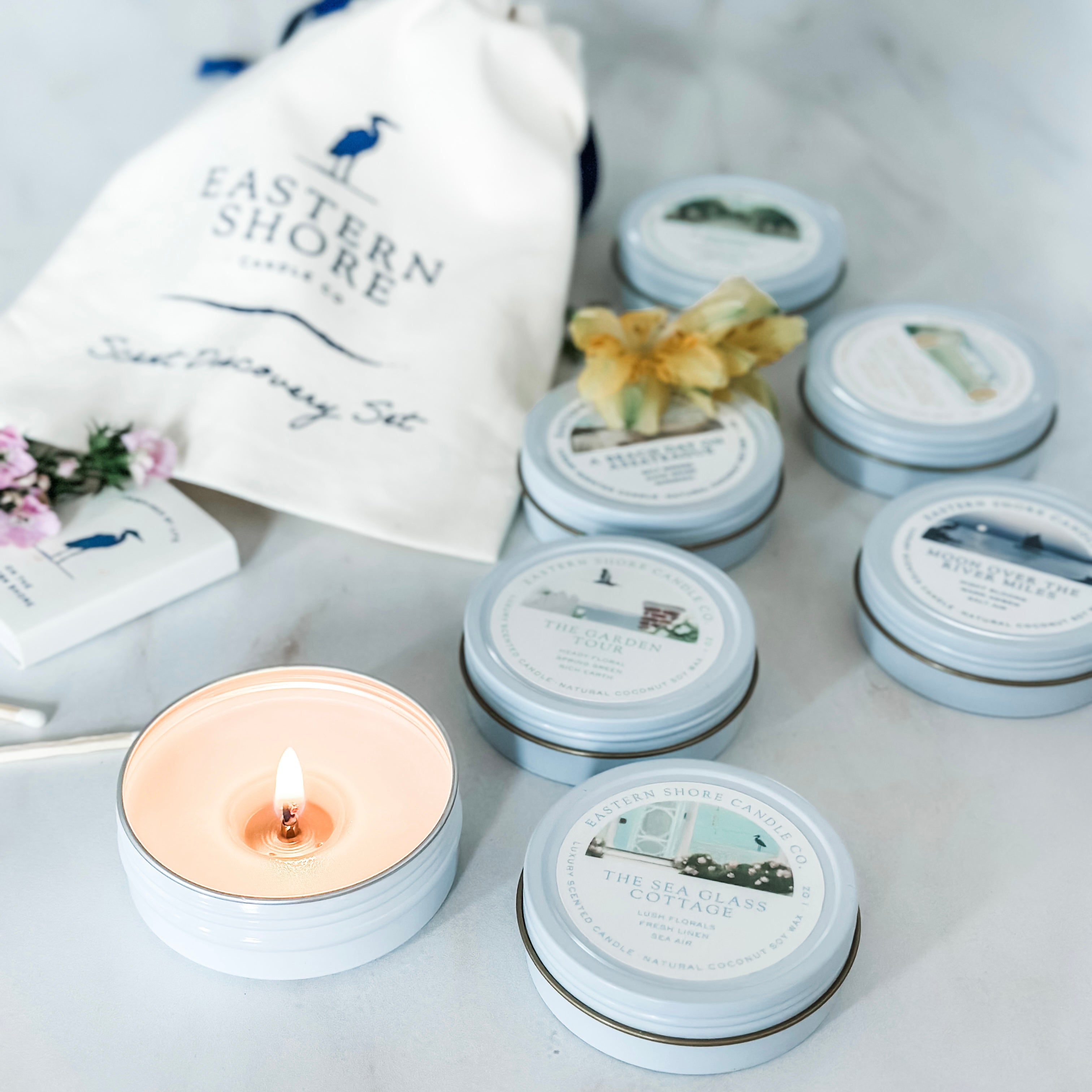 A collection of tin candles with various labels, arranged around a lit candle, with a heron wooden match used for lighting. A fabric bag with the text &