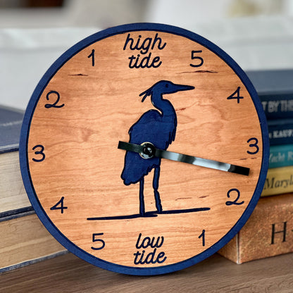 A round wooden wall clock with a navy blue border, a heron silhouette, and tide-themed text reading &