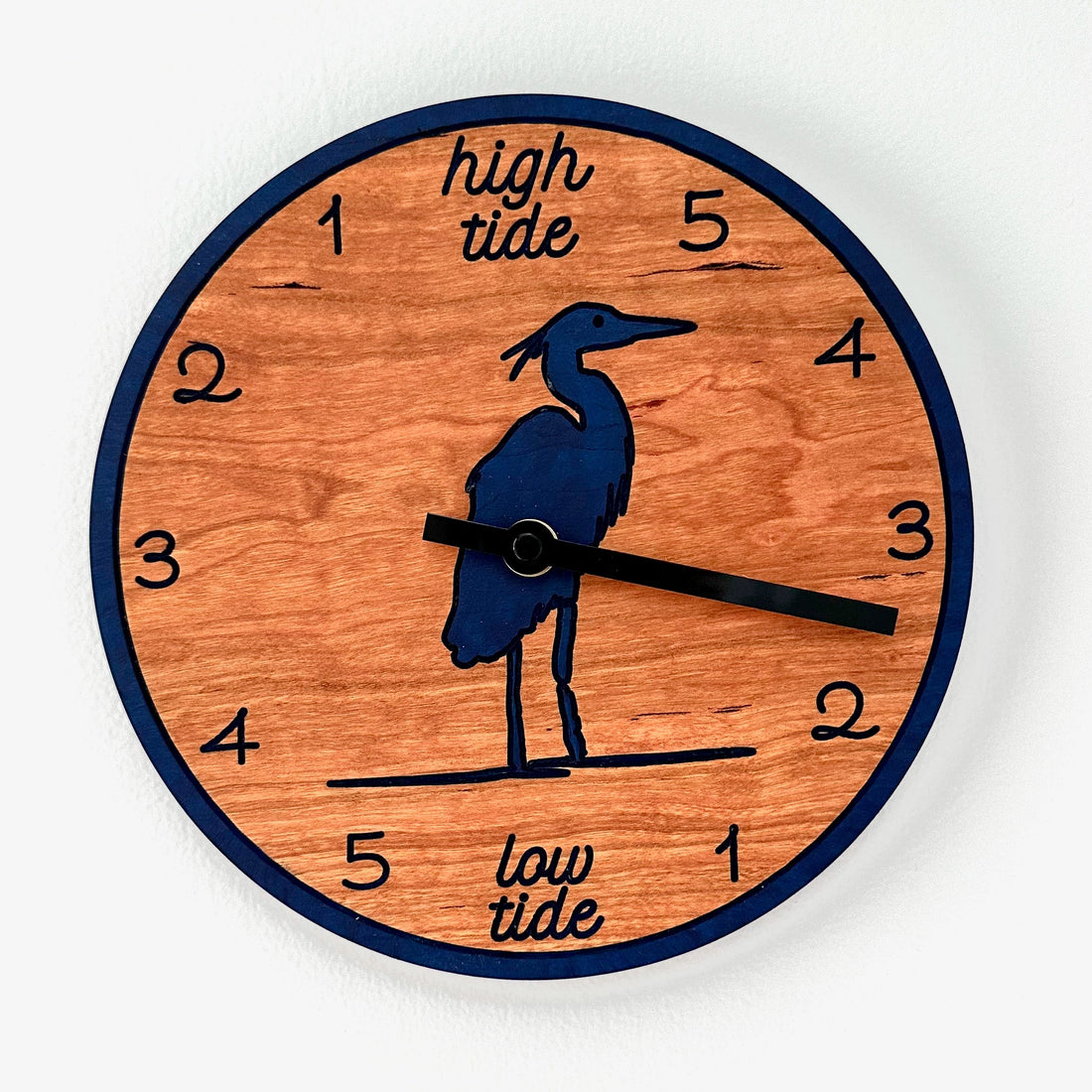 A round wooden wall clock with a dark blue border, navy blue heron silhouette, and black clock hands. The clock face features black numerals and the words &